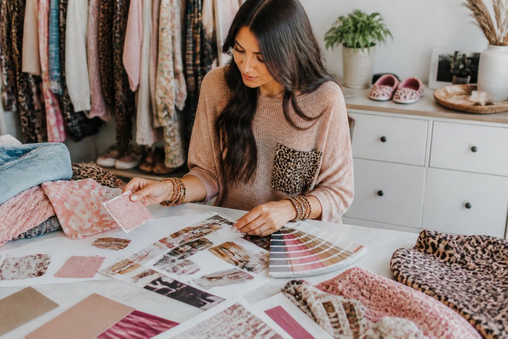 Fashion-forward woman shopping online on her laptop, surrounded by fabric swatches, color palette boards, and style inspiration clippings on a bright workspace