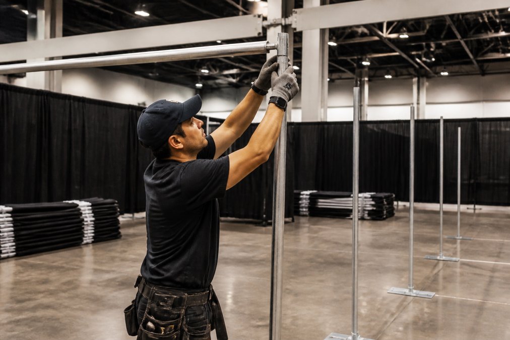 Close-up of technician securing crossbar to telescopic uprights during installation, demonstrating proper pipe and drape assembly with safety equipment visible and base plates anchored