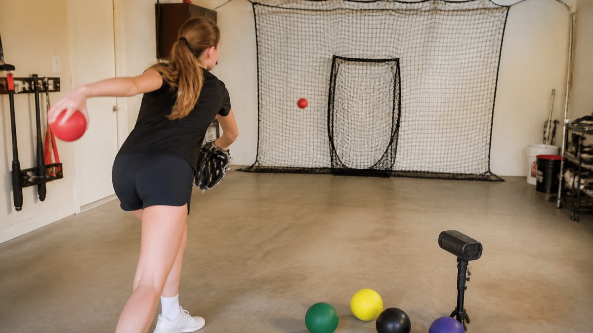 A young female fastpitch softball pitcher throwing a weighted ball into a target net in a garage or indoor training space, with a radar gun and several colored plyo balls on the floor nearby