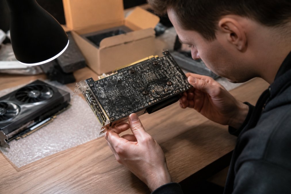 A person inspecting a second-hand graphics card closely under a bright desk lamp, examining the PCB and connectors carefully for signs of damage or burn marks