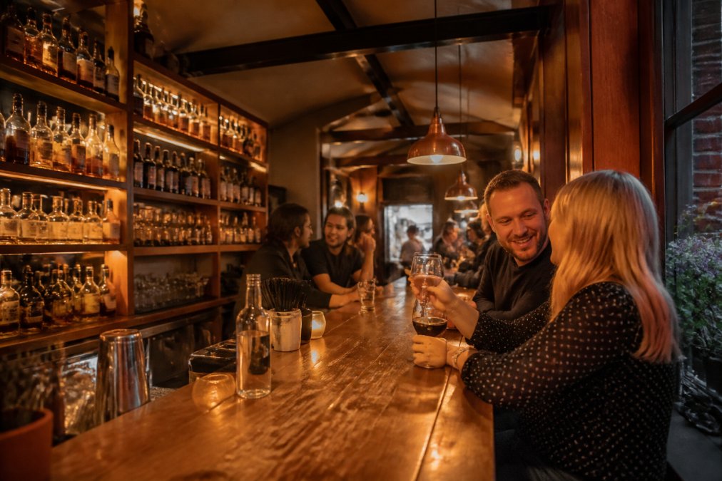 Warm interior of Plonkers Wine Bar with a wooden bar lined with amber spirit bottles, a few patrons chatting over drinks in soft evening light