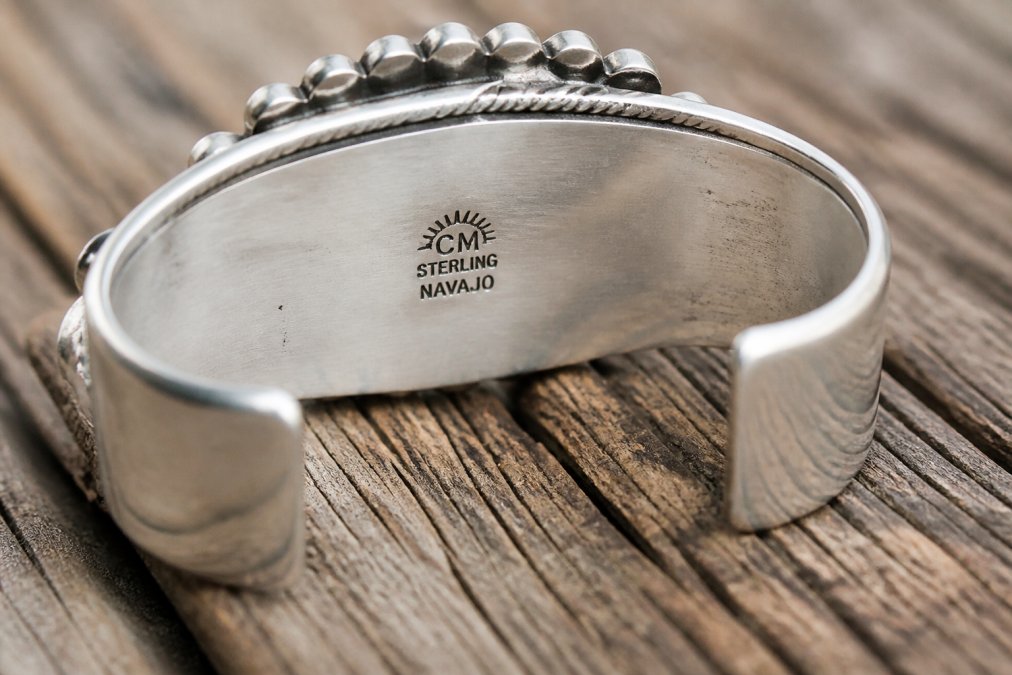 Close-up of the underside of a heavy sterling silver Navajo cuff bracelet resting on a weathered wooden surface, showing a clear hallmark stamp with artisan initials, a sterling mark, and tribal notation, photographed in warm natural light