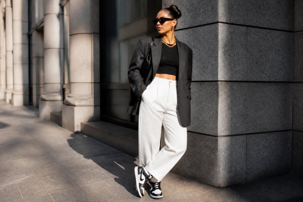 Confident woman in her 30s wearing a bold streetstyle outfit with oversized blazer, statement chain necklace, and high-top sneakers, posing against modern urban architecture with morning sunlight casting dramatic shadows