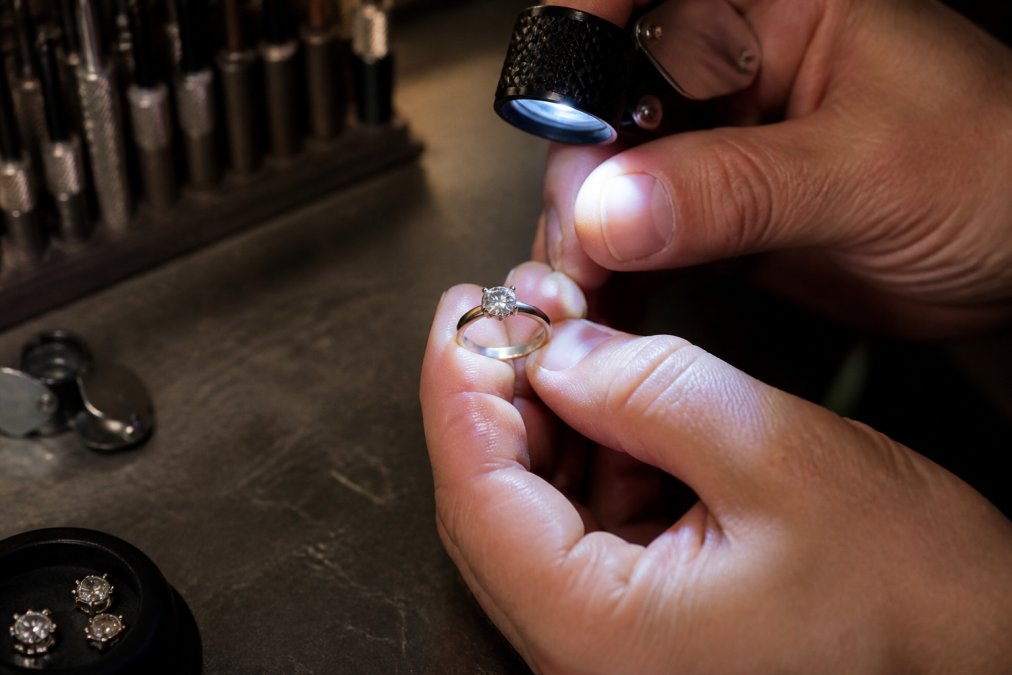 A close-up of a professional jeweller's hand holding a diamond ring under a bright loupe light on a dark workshop bench, examining the stone's facets