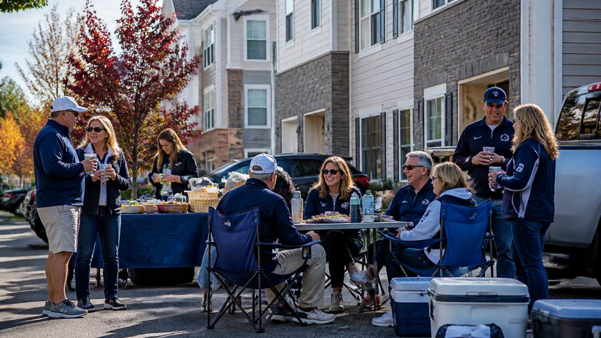 A group of alumni in blue and white attire tailgating outside a modern townhome near a college campus on a sunny fall game day, with coolers and folding chairs visible
