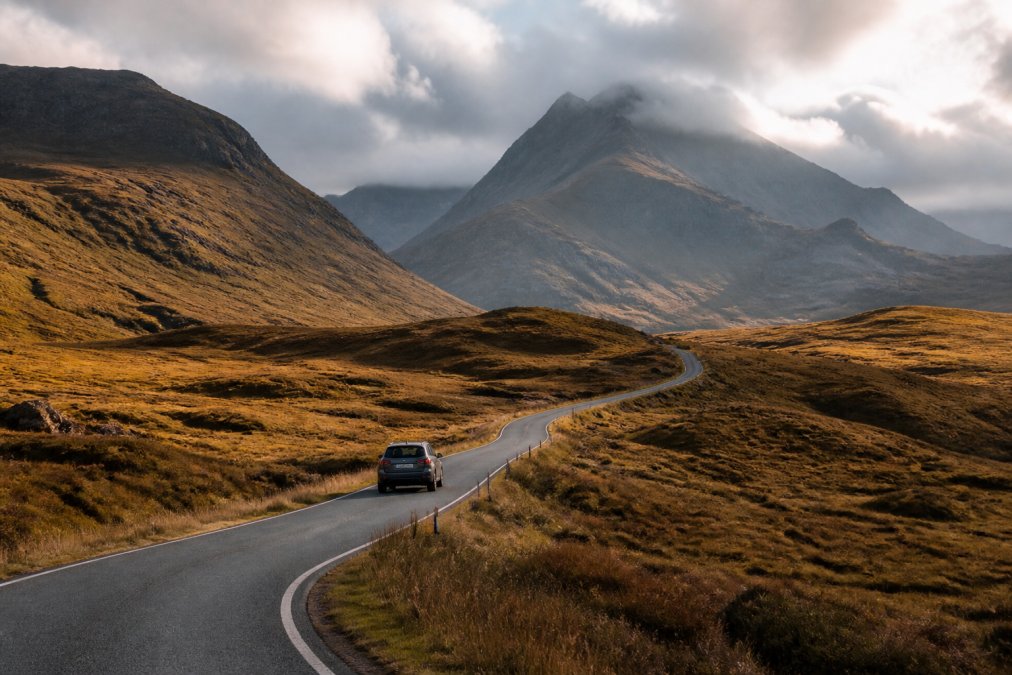 A winding Highland road stretching into dramatic mountain landscape with a single car navigating the route, misty peaks in distance, late afternoon light casting long shadows across the terrain