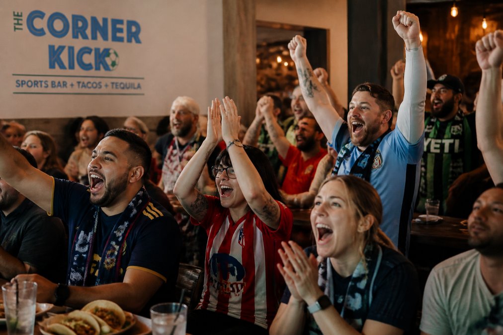 Diverse group of soccer fans gathered indoors wearing colorful team jerseys and scarves, celebrating a goal with raised arms and genuine enthusiasm, with varied expressions of excitement and camaraderie