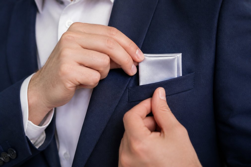 Close-up of hands demonstrating the presidential fold technique on a silk pocket square, showing precise alignment and the neat rectangular point emerging from a jacket pocket with natural lighting