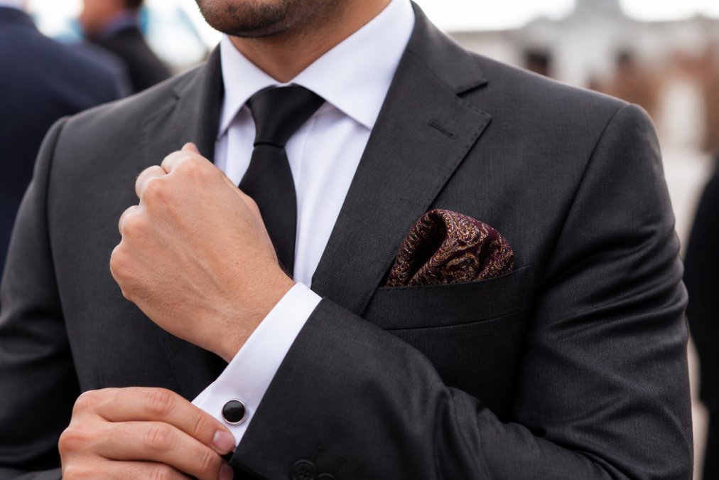 Groom in charcoal suit jacket showing coordinated black silk tie with complementary pocket square featuring subtle paisley pattern in burgundy and gold tones, with matching cufflinks visible
