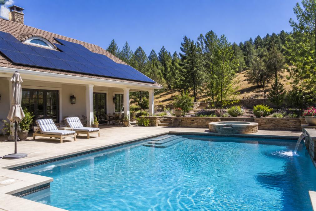 Residential swimming pool with solar panels mounted on roof in bright sunlight, showing integration with pool deck and surrounding landscape