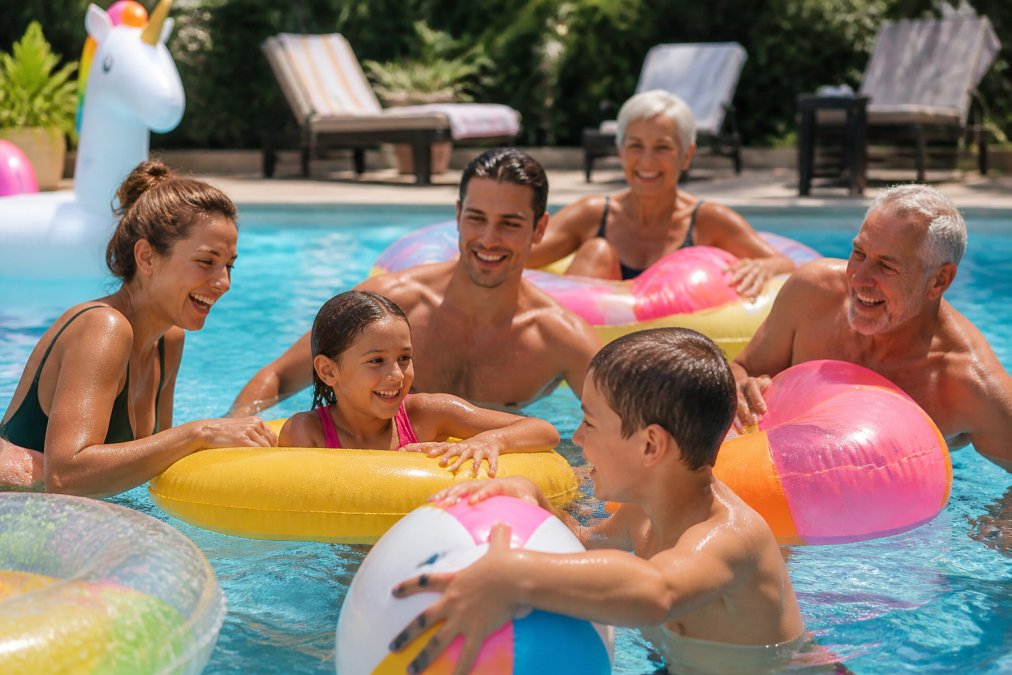 A multi-generational family laughing together in a sparkling pool, children and adults engaged with colorful inflatable toys and games, bright afternoon sunlight reflecting off the water