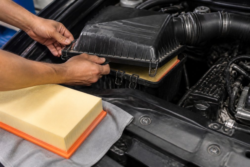 A pair of hands unlatching the spring-steel housing clips on a Porsche Cayenne airbox assembly in a well-lit engine bay, with the upper lid partially open revealing the old paper element filter inside, a new OEM filter resting on a clean shop cloth nearby
