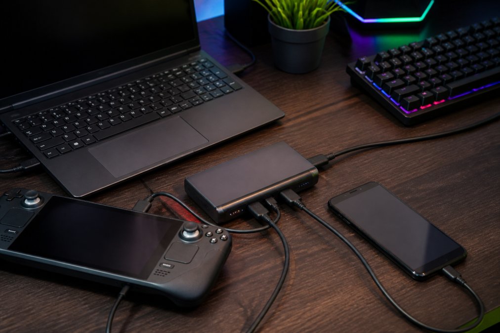 Gaming setup with laptop, Steam Deck, and smartphone arranged around a portable charger on a desk with RGB lighting and gaming peripherals visible