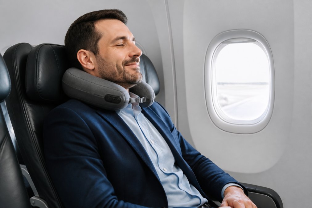 Business traveler using a cordless neck massager while seated in airplane cabin with proper posture, demonstrating comfortable usage during flight
