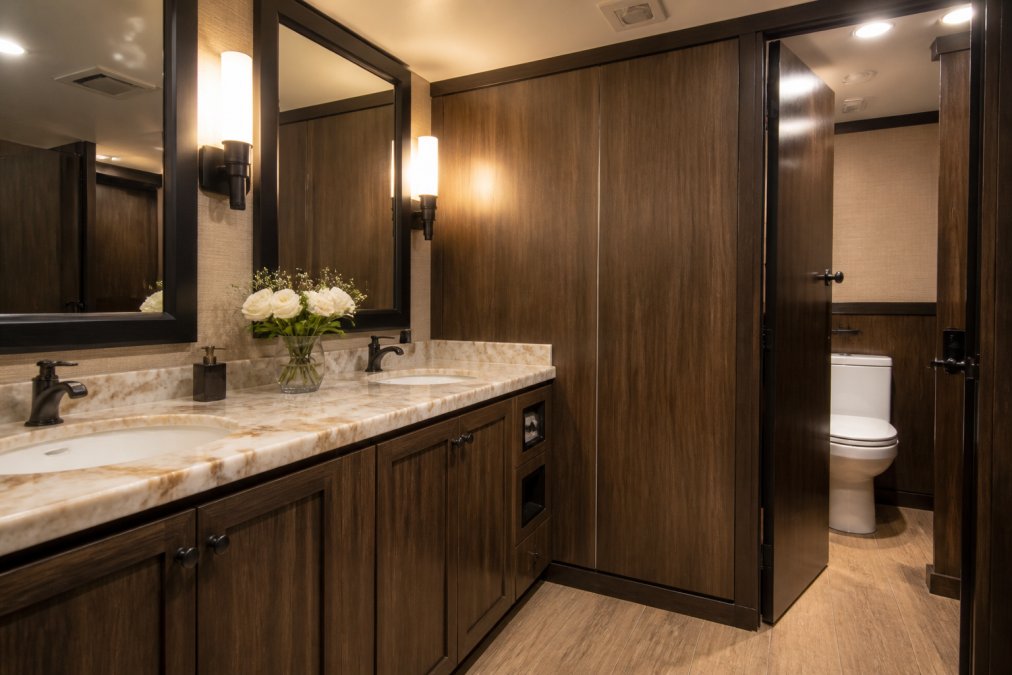 Interior of an upscale restroom trailer with warm interior lighting, vanity mirrors above a marble countertop, fresh flowers in a small vase, wood-paneled walls, and a flushing toilet visible through a partially open stall door