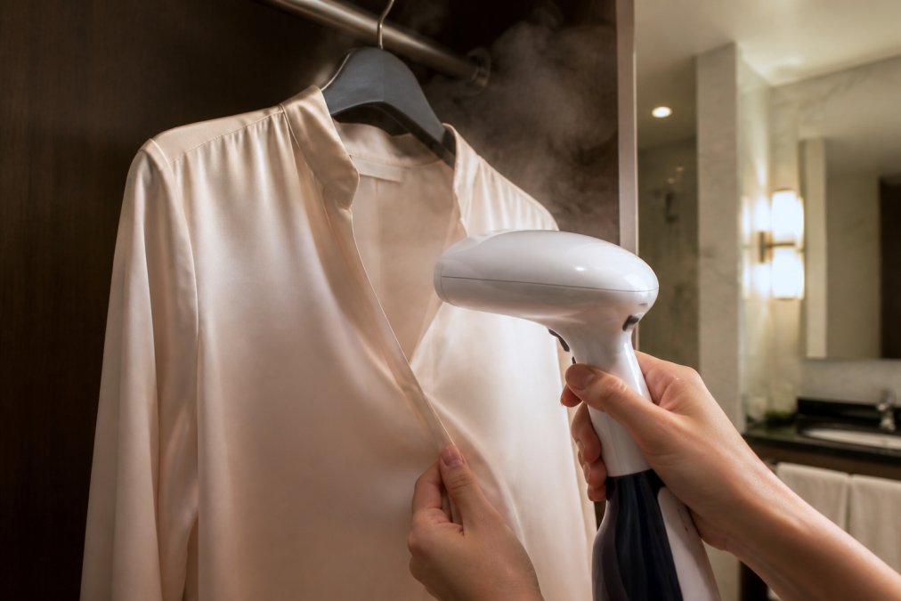 Close-up of hands holding a compact handheld garment steamer against a silk blouse hanging on a hotel closet rod, steam visibly rising, warm bathroom lighting