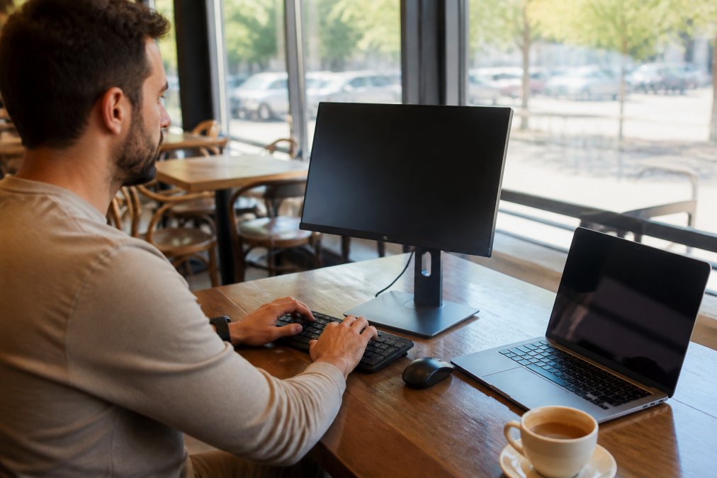 Digital nomad working at a café with a portable OLED monitor elevated on a stand, wireless keyboard and mouse positioned on the table, laptop to the side, natural daylight streaming through café windows, coffee cup visible on the corner of the desk