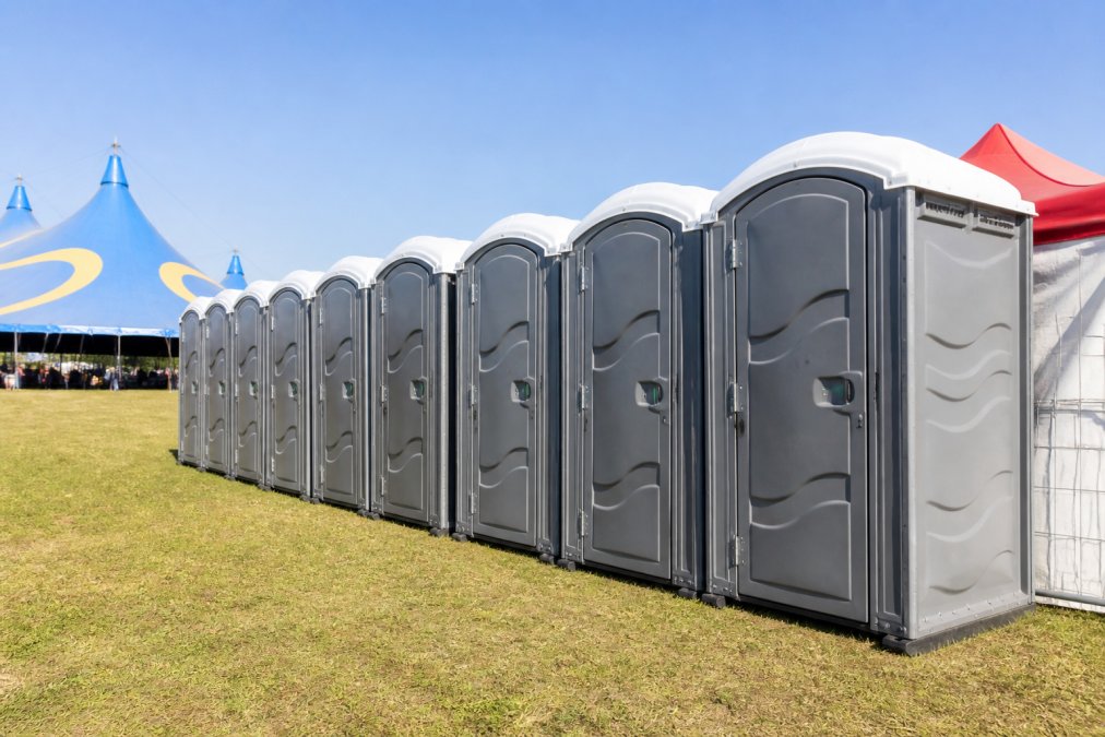 A row of clean, modern portable restroom units set up on grass at an outdoor festival, clear blue sky overhead, colorful event tents in the background, morning light casting soft shadows