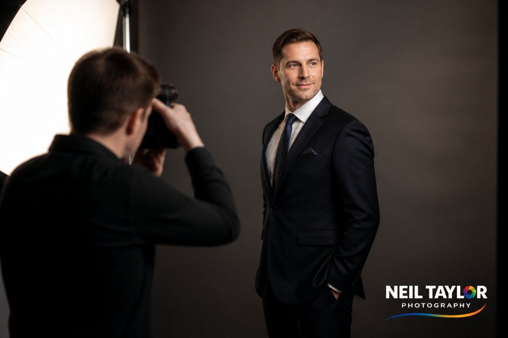 Professional in tailored business suit demonstrating angled stance with shoulders at 45 degrees to camera, chin slightly lifted, standing against neutral grey studio background with soft directional lighting