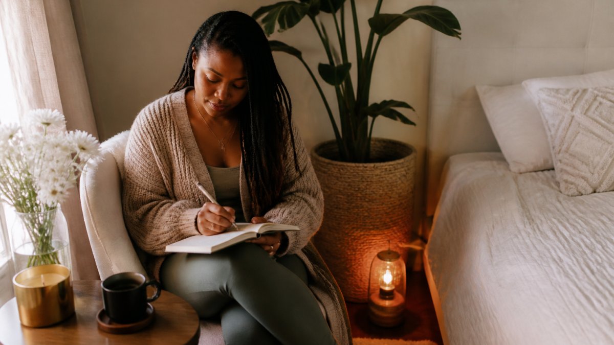 Woman sitting peacefully in a sunlit bedroom corner journaling with a warm cup of tea