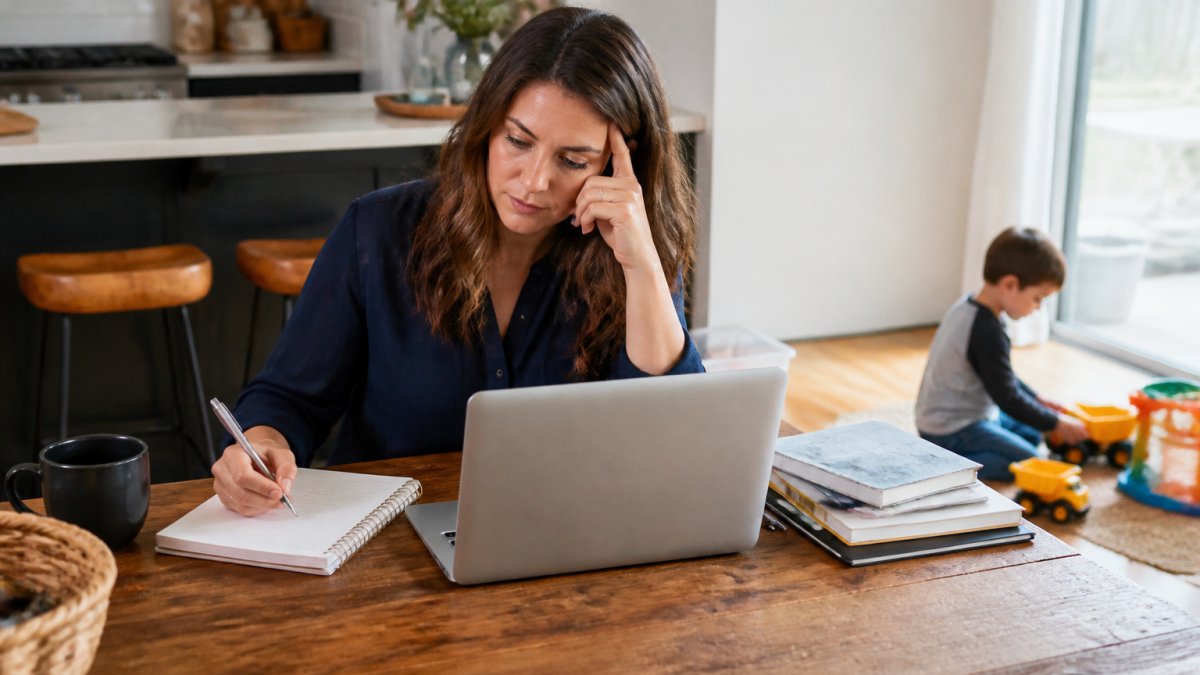 A parent at a kitchen table with a laptop open, a notebook and coffee beside them, working in a quiet moment while a child plays nearby in soft focus