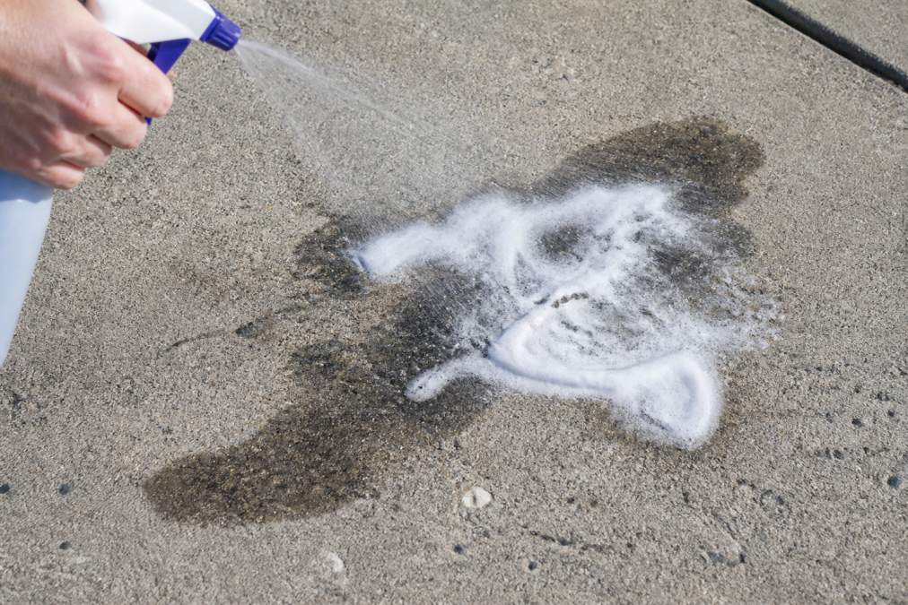 Close-up of a grey concrete driveway surface showing a dark, irregular oil stain being treated with white degreaser foam sprayed from a bottle, foam pooling on the textured concrete surface in bright afternoon sunlight