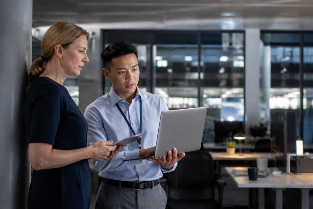A risk management officer and a security architect reviewing compliance dashboards on large monitors in a modern office, audit trail logs visible on screen, cool overhead lighting