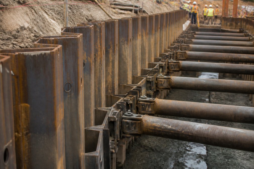 Close-up of interlocking steel sheet piles and hydraulic shore props installed in an active deep excavation, workers in safety gear visible in background, daylight showing the mechanical precision of the shoring installation