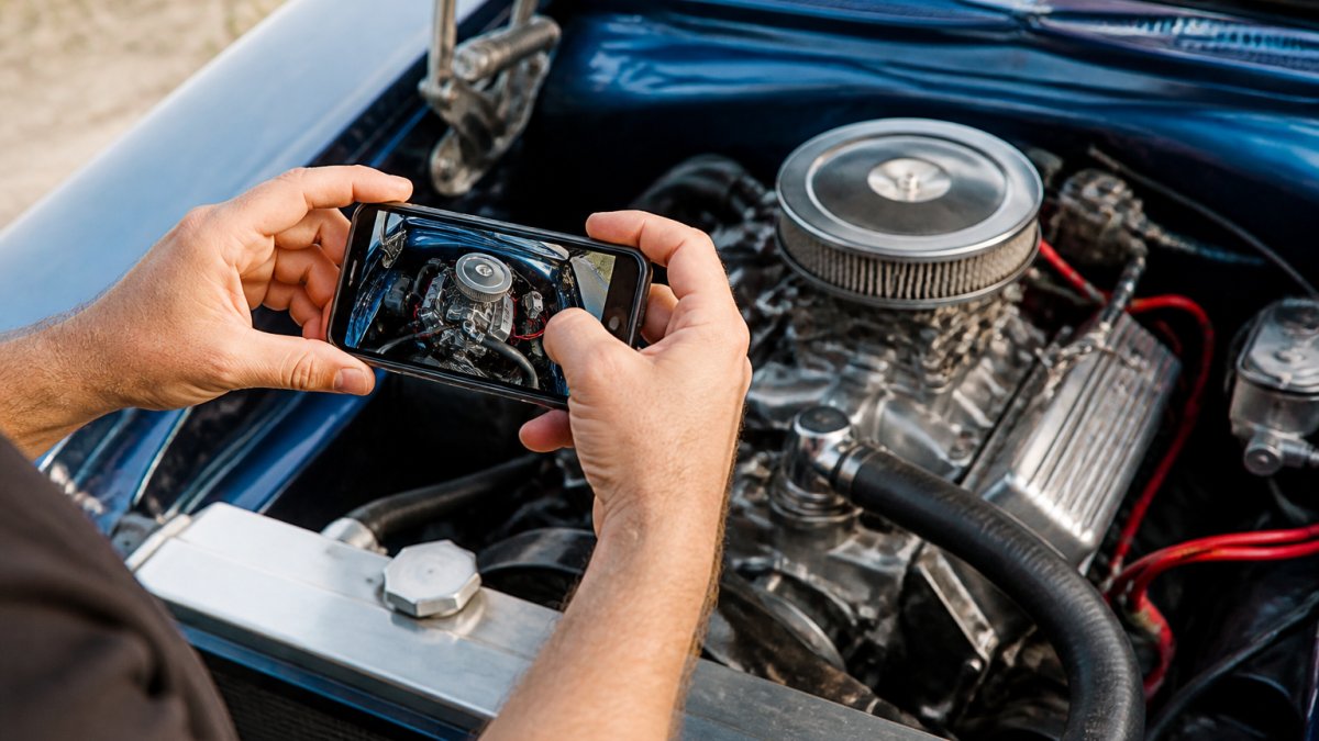 Hands holding a smartphone photographing an exposed engine bay of a classic car, showing before-and-after angles with natural daylight illuminating mechanical details