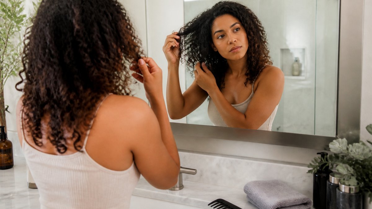 Woman with curly hair checking her texture in a mirror during hair maintenance with a comb and towel nearby