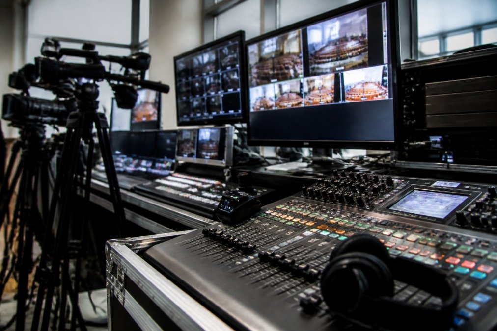 Professional broadcast control room with multiple monitors displaying video feeds, audio mixing console with faders, camera equipment, and headphones during active AGM production