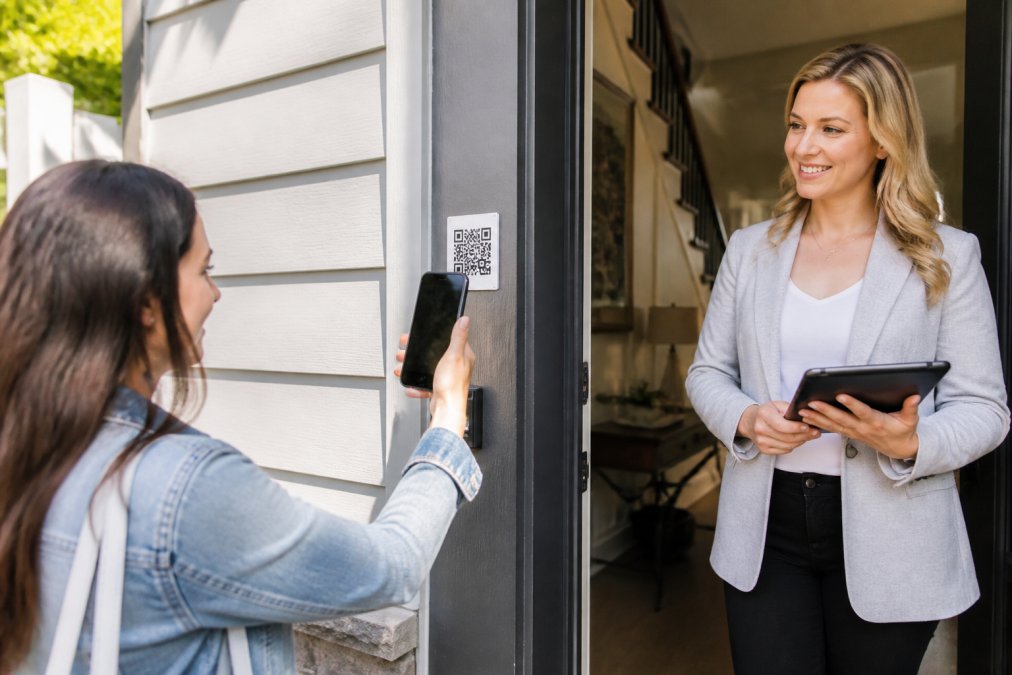 Real estate agent holding tablet at property entrance with QR code visible on doorframe, visitor scanning code with smartphone in afternoon sunlight
