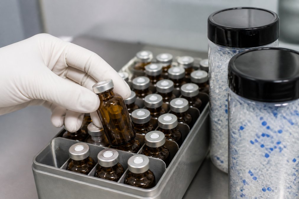 Close-up of amber glass vials with PTFE-lined caps arranged in storage rack, protective labelling visible, desiccant canisters positioned nearby, white gloved hand holding one vial