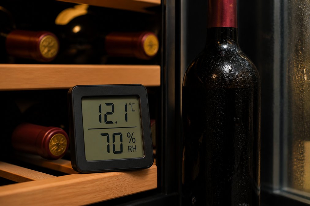 A close-up of a digital hygrometer and thermometer inside a dark, temperature-controlled wine cabinet, with a bottle of red wine visible in the background, showing condensation on the glass