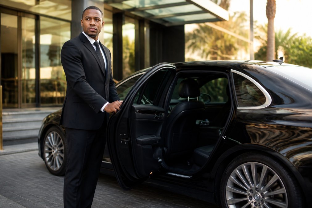 A professionally dressed chauffeur in a dark suit holding open the rear door of a polished black luxury sedan, standing at attention with a neutral, composed expression outside a glass-fronted hotel entrance under soft morning light