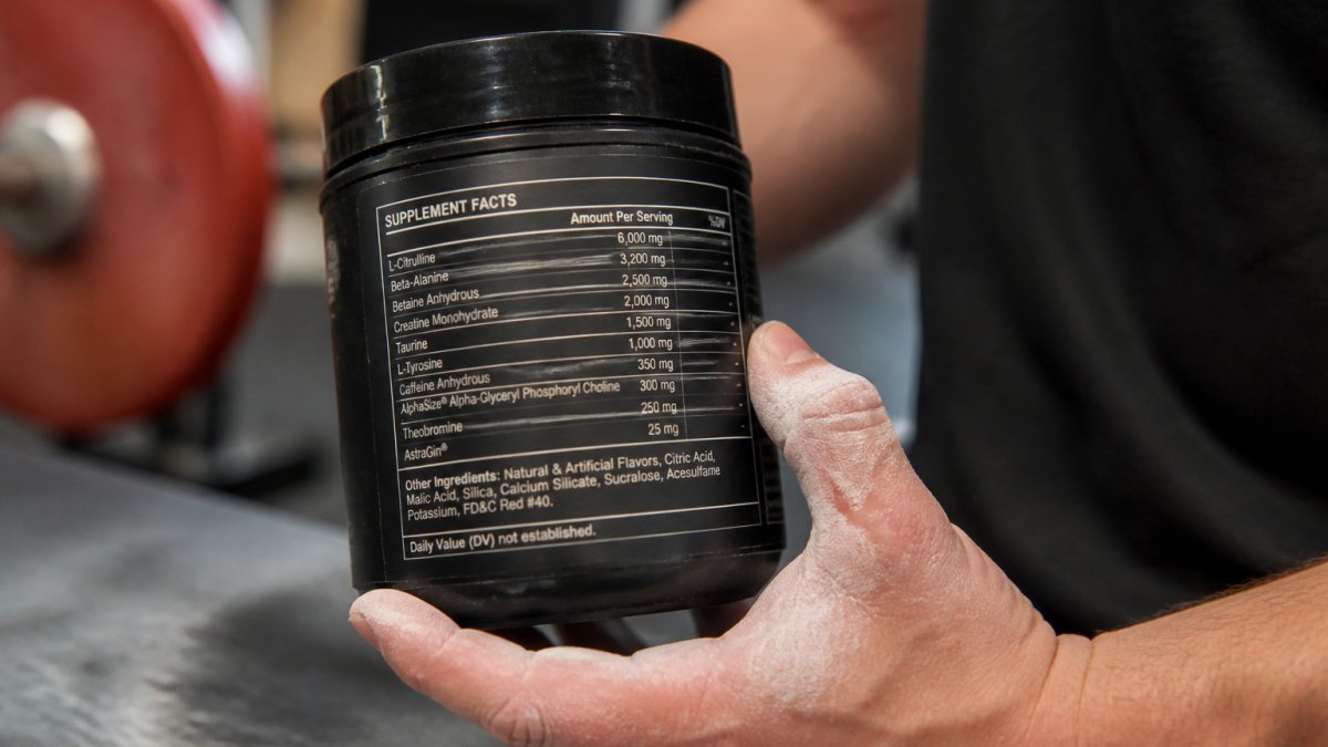 Close-up of a pre-workout supplement label showing ingredient doses in milligrams and grams, with a lifter's chalked hand holding the tub