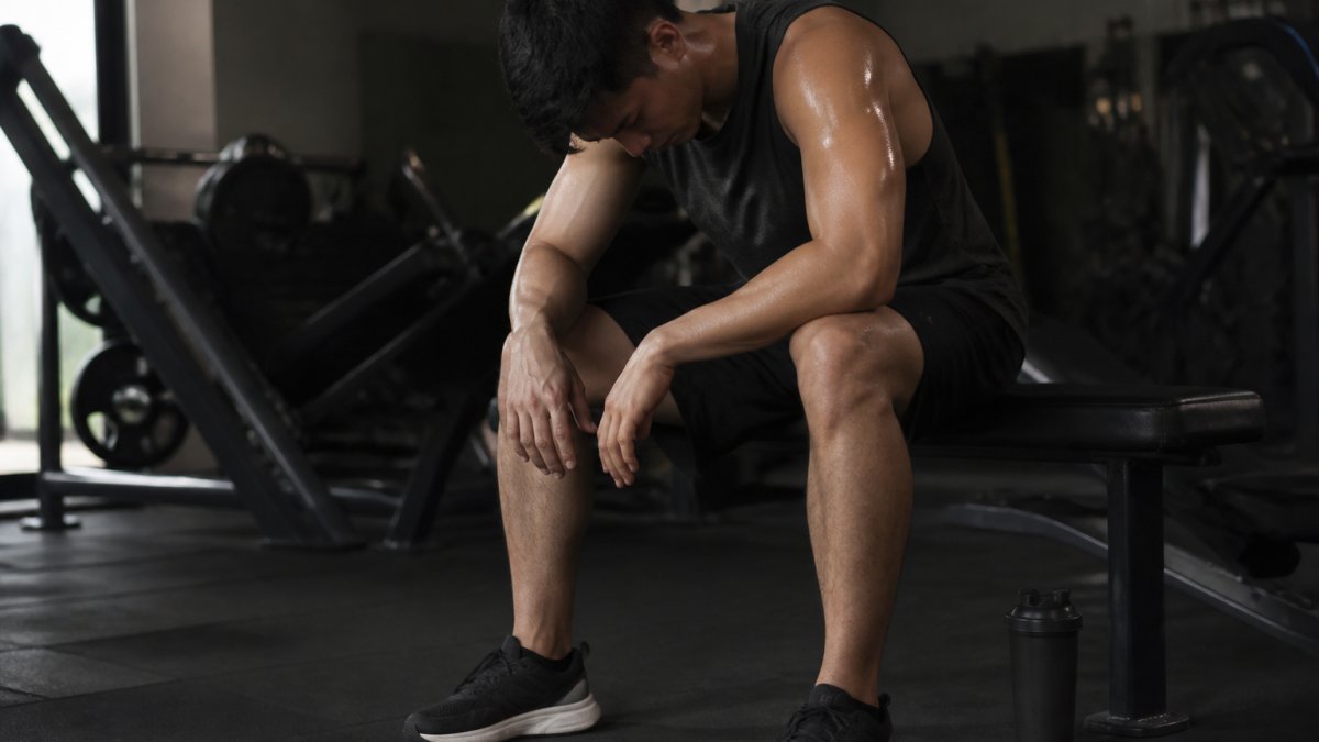 A tired gym-goer sitting slumped on a bench in a dimly lit gym, sweat visible, a shaker bottle resting on the floor beside them