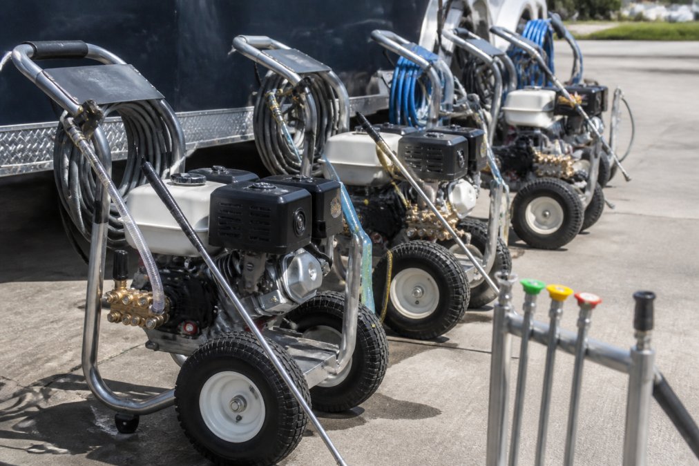 Commercial pressure washing equipment lined up on a job site, showing gas-powered units with visible hoses, quick-connect nozzles, and aluminum frames positioned ready for deployment on concrete surfaces