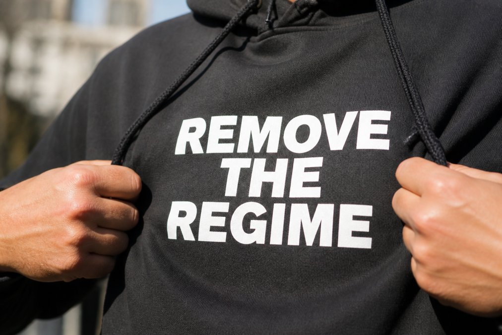 A close-up of a person's hands gripping the fabric of a black hoodie, with a bold white political slogan printed across the chest showing crisp, sharp edges under natural daylight