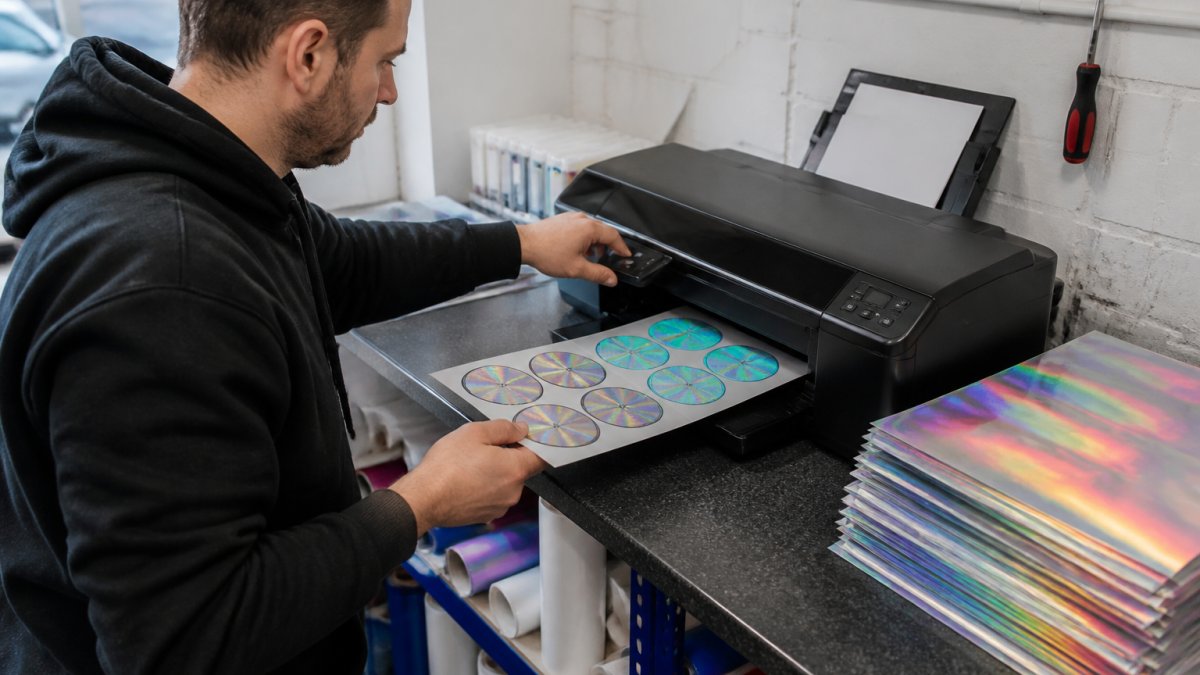 A print operator in a small UK sign shop adjusting printer settings on a desktop inkjet, with sheets of rainbow holographic sticker paper stacked beside the machine and a test print showing a clean, smudge-free sticker design