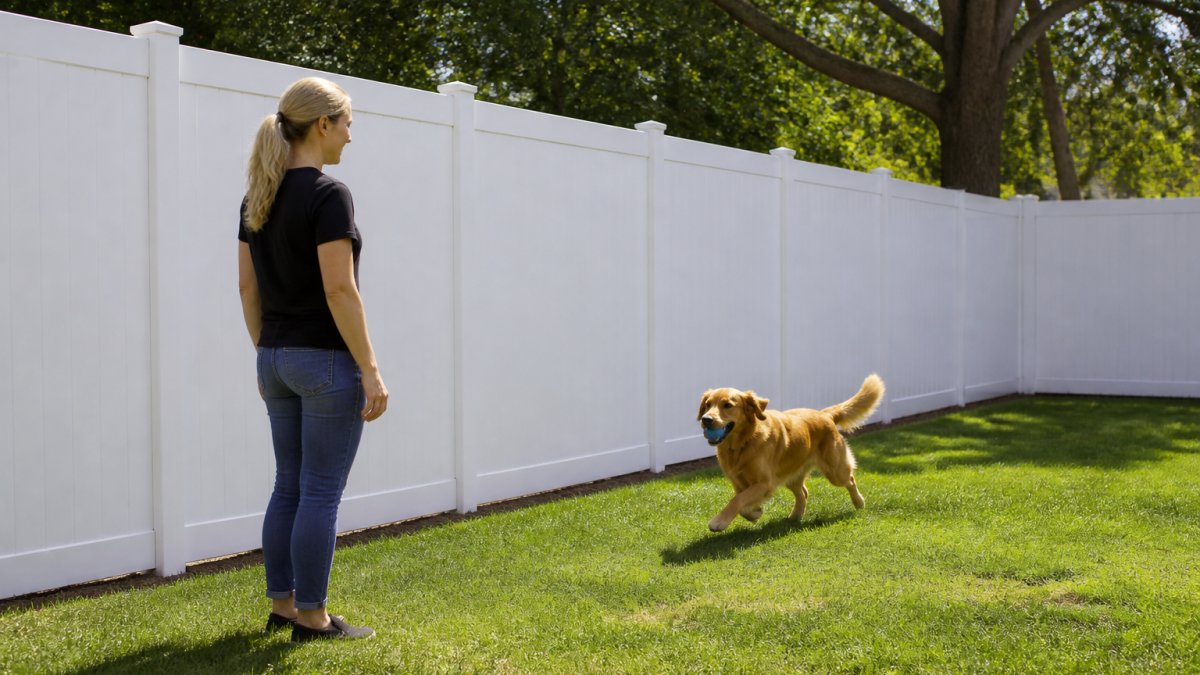 Homeowner standing beside newly installed vinyl privacy fence with dog playing safely in enclosed yard, showing solid white panels and ground-level closure preventing escape, natural afternoon sunlight