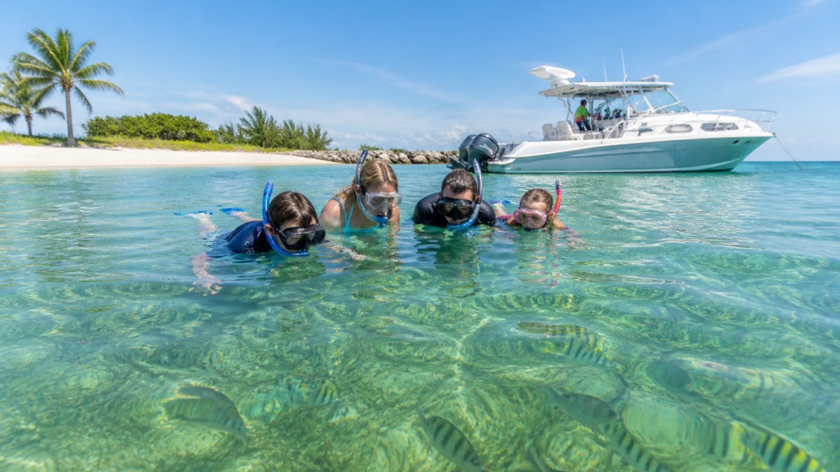 A family in snorkel gear swimming in clear shallow water near a sandy island shoreline, tropical fish visible below the surface, a private boat anchored in the background