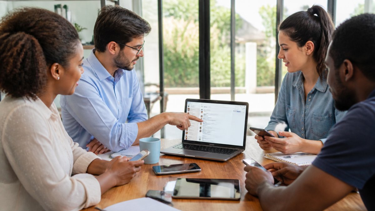 A small business team of four people collaborating around a table with laptops and smartphones, one person pointing at a screen showing a chat interface, bright modern office with large windows