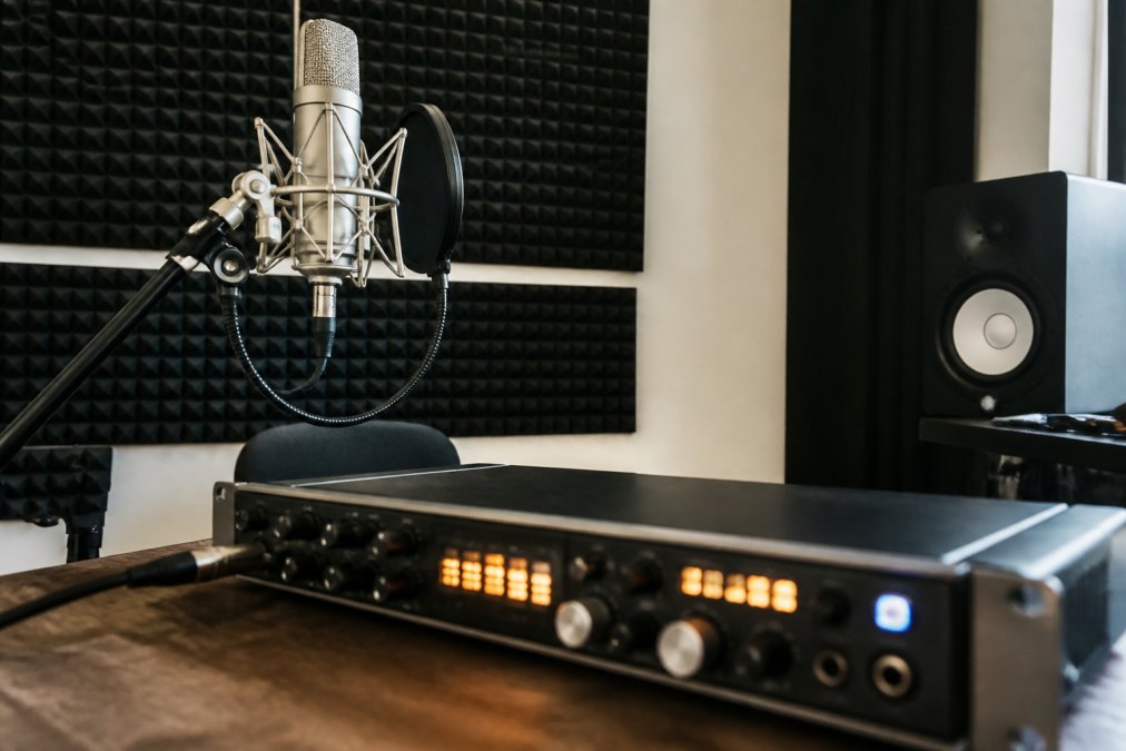A large-diaphragm condenser microphone mounted on a boom stand inside a home studio, with acoustic foam panels visible on the wall behind it and an audio interface with glowing input meters in the foreground
