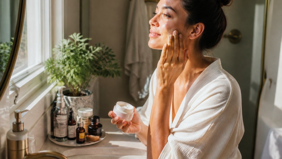 Person in morning light applying skincare products at bathroom vanity with natural sunlight, showing a calm, intentional morning ritual with beauty products arranged nearby