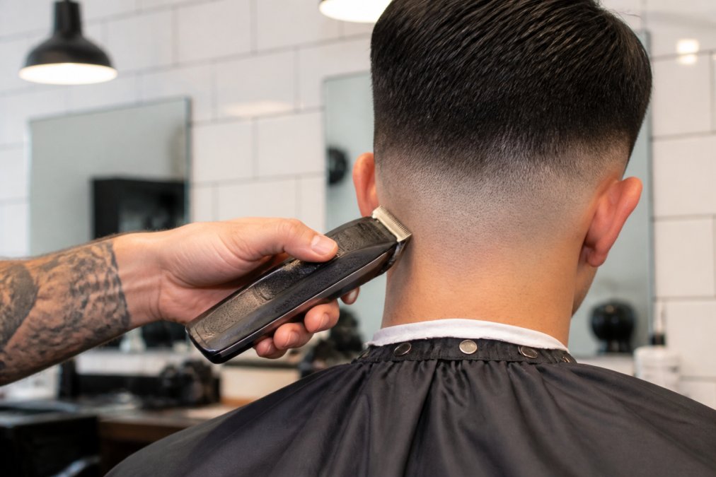 Close-up of a professional barber's right hand gripping a cordless clipper at the nape of a client's neck, performing a skin fade in a bright, clean barbershop with white tile walls and warm overhead lighting
