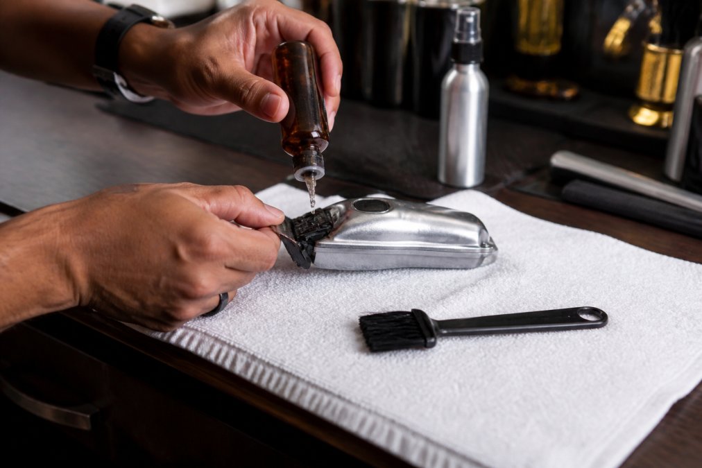 Barber's hands applying two drops of blade oil from a small amber bottle onto the open teeth of a professional clipper laid flat on a white station towel, with a cleaning brush and blade spray visible in the background on a wooden barbershop counter