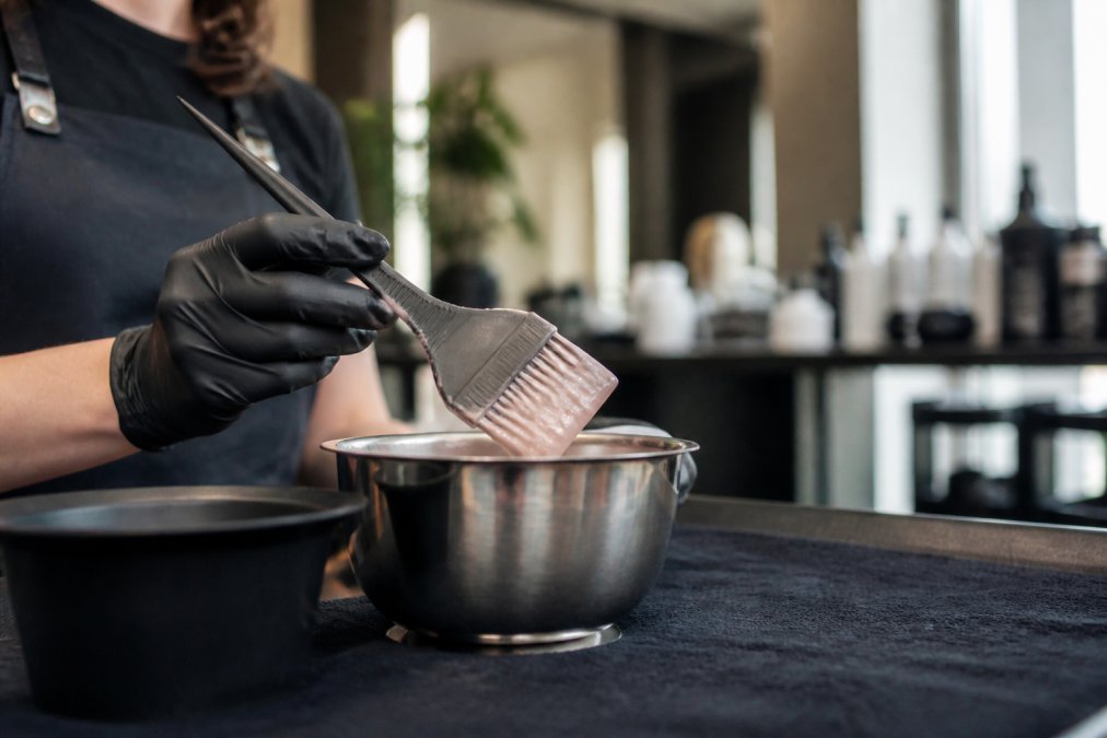 Close-up of a professional colorist's hand holding a feathered tint brush over a stainless mixing bowl in a modern salon studio, dye visible on the brush tip, soft window lighting