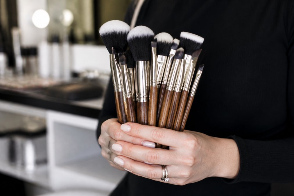 Professional makeup artist's hands holding a collection of Girl Meets Brush brushes with various bristle sizes arranged by type, soft studio lighting illuminating the synthetic bristles and wooden handles