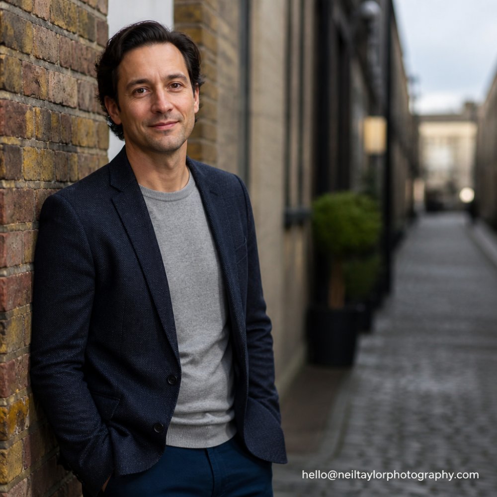 An actor in a casual blazer standing by a brick wall in soft, overcast daylight, looking relaxed and approachable, with a shallow depth of field blurring the background
